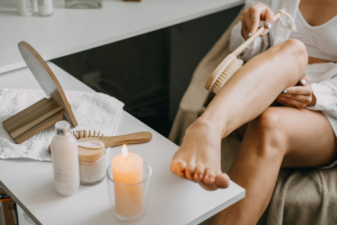 Dry brushing body brush for exfoliating dry skin, lymphatic drainage and cellulite treatment. Woman brushing skin leg with dry wooden brush to prevent cellulite and body problem in bedroom at home